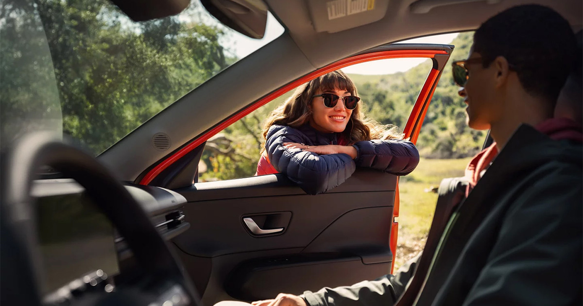A woman with sunglasses on smiling and leaning through the window of a vehicle's open passenger door at the driver.